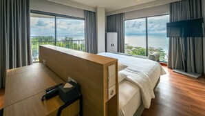 Habitación de hotel con cama, escritorio, televisión y ventanas amplias con vista al mar.