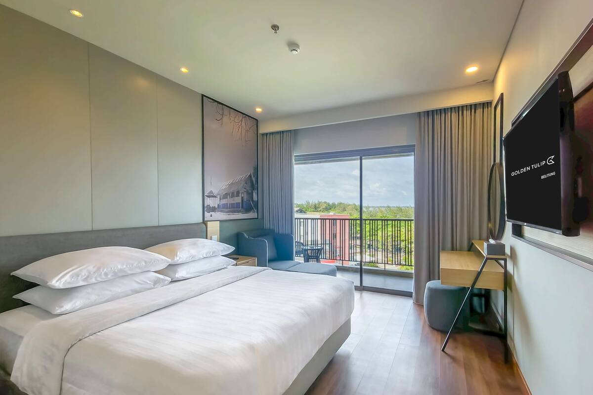 A hotel room with a large bed, a flat-screen TV, a balcony with a view, and a painting on the wall.