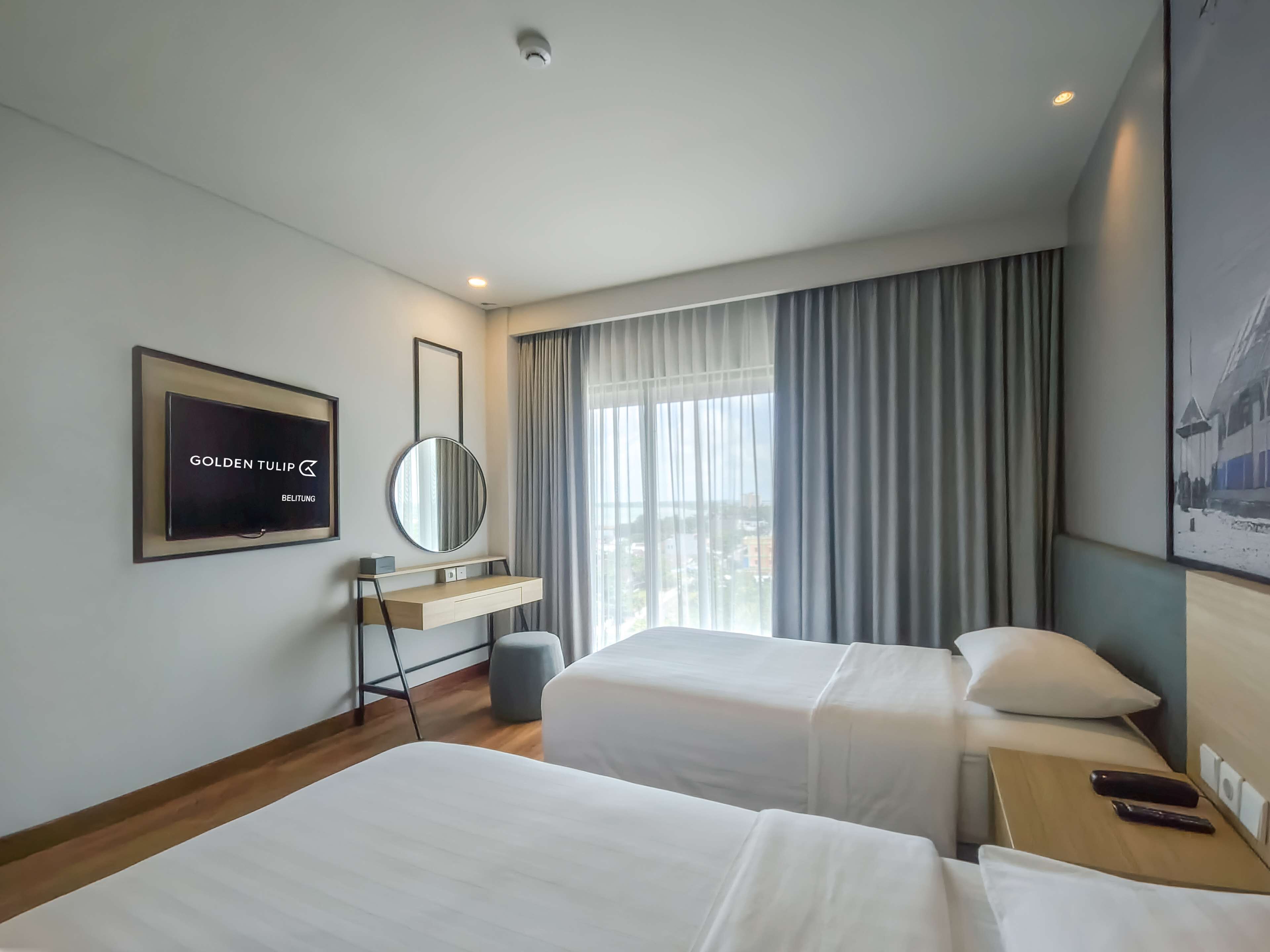 A hotel room with two beds, a TV, a mirror, and a window with curtains.
