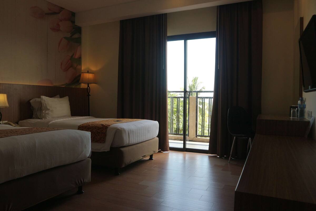 A hotel room with two beds, a desk, and a balcony with curtains.