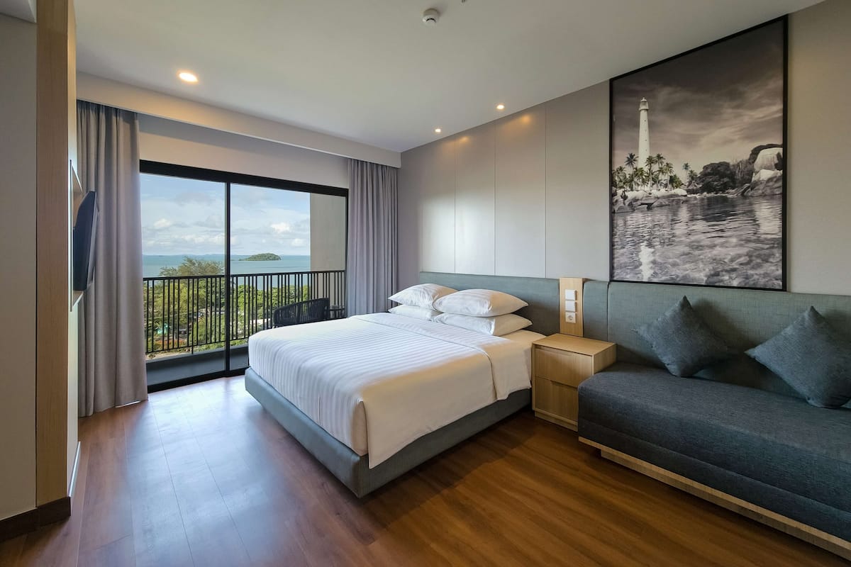 A modern hotel room with a large bed, a sofa, a painting of a coastal scene, and a balcony with a view of the sea.
