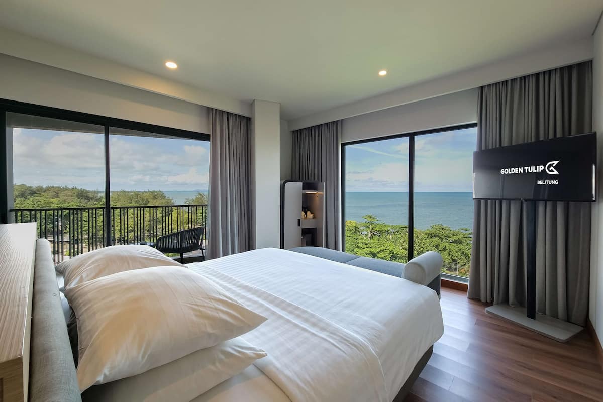 A hotel room with a large bed, a TV on the wall, and a balcony with a view of the sea.