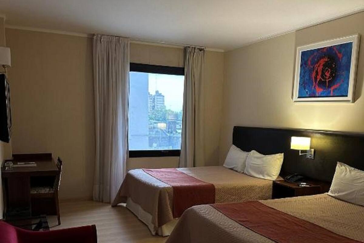A hotel room with two beds, a desk, a chair, a window with curtains, and a painting on the wall.