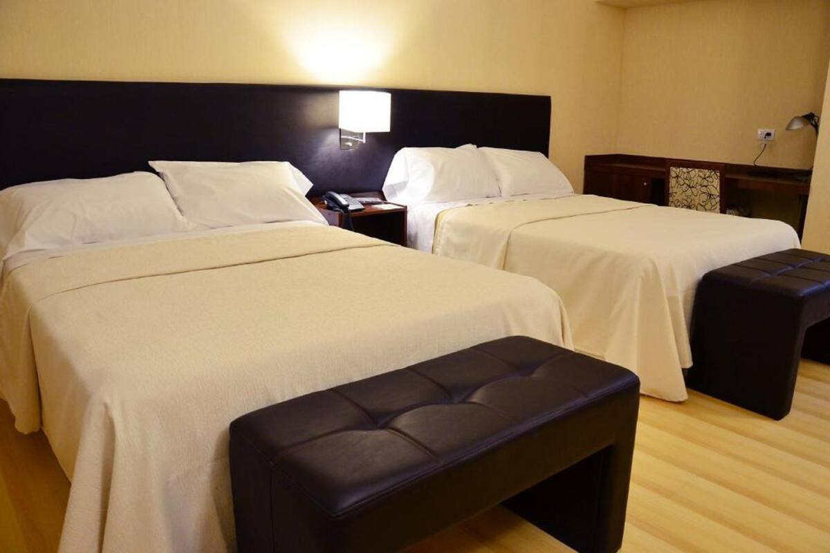 A hotel room with two beds, a bench, a nightstand with a lamp, and a small table with a phone.