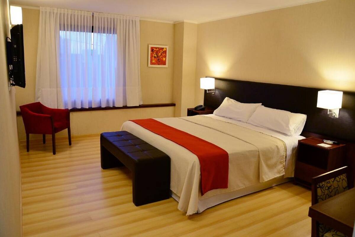 A hotel room with a bed, a red chair, a bench, a nightstand with a lamp, and a window with curtains.