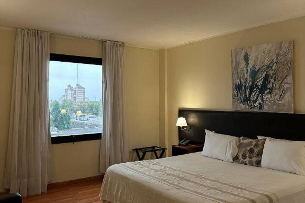 A hotel room with a large bed, a nightstand, a lamp, a painting on the wall, and a window with curtains.