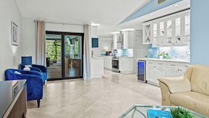 A modern living room with a glass coffee table, a beige sofa, and a blue armchair. A kitchen with white cabinets and a blue countertop is visible in the background. - Villa Turquoise Coast (Cape Coral)