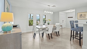 A modern kitchen with a dining area, featuring a light-colored countertop, white chairs, and a blue patterned bar stool. - Villa Turquoise Coast (Cape Coral)