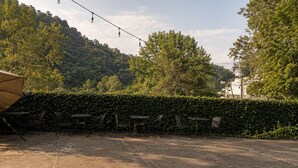 Un espace extérieur aménagé en terrasse, avec des tables et des chaises, entouré d’une haie et d’arbres.