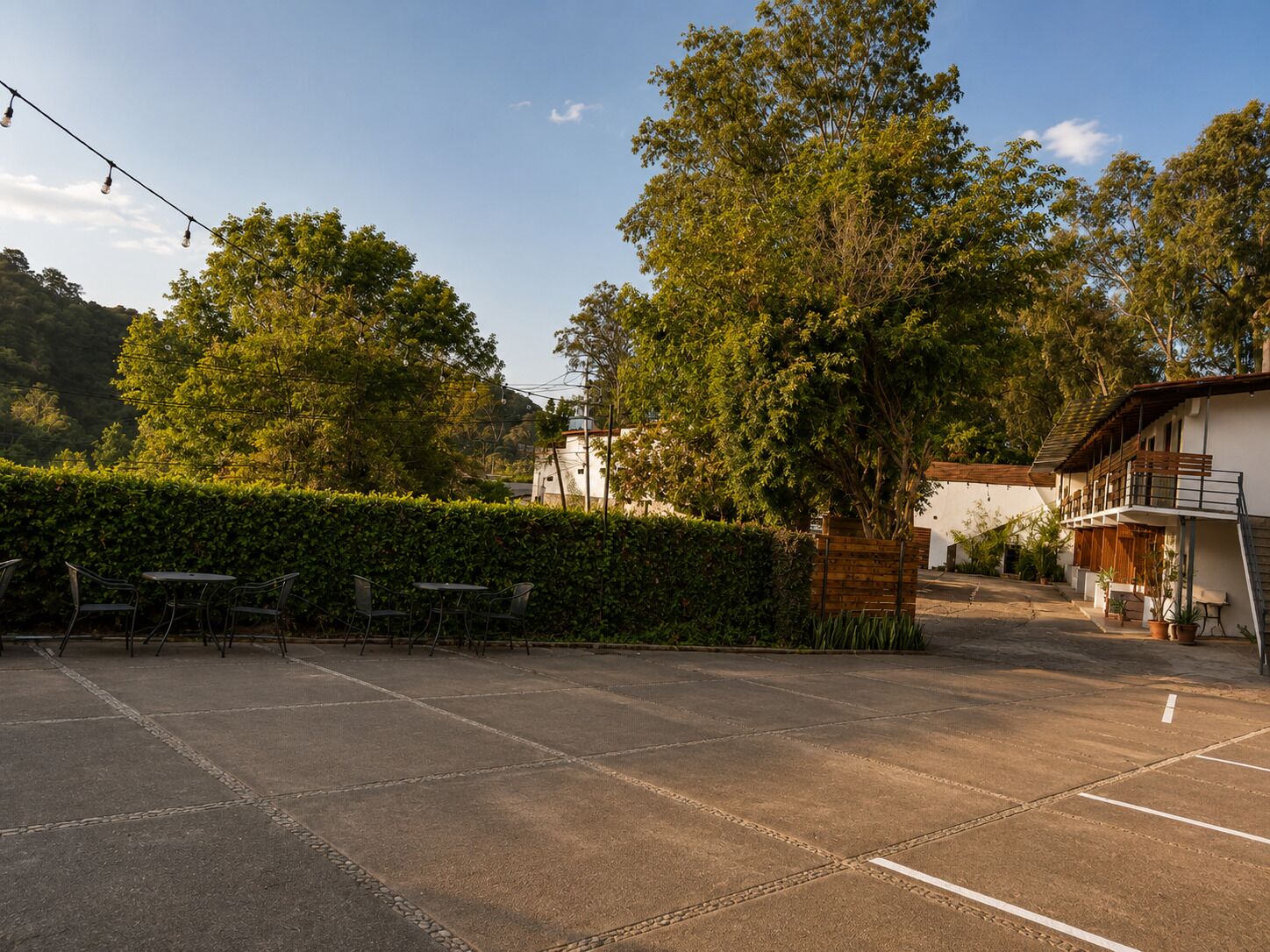 An outdoor seating area with tables and chairs, surrounded by a hedge and trees.