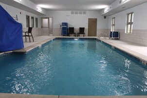 Una piscina interior con agua cristalina, paredes y suelo azulejados. Se observan sillas y una zona techada en un lateral.