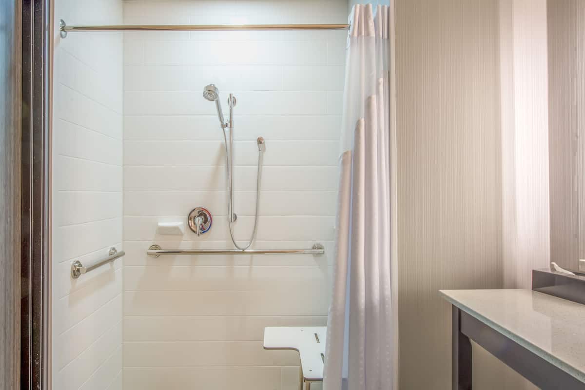 Un baño moderno con paredes de azulejos blancos, una ducha con cabezal de ducha manual, un inodoro y un lavabo.
