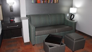 Habitació d'hotel amb un sofà, una taula petita amb una làmpada, una taula auxiliar, un moble amb calaixos i un quadre a la paret.