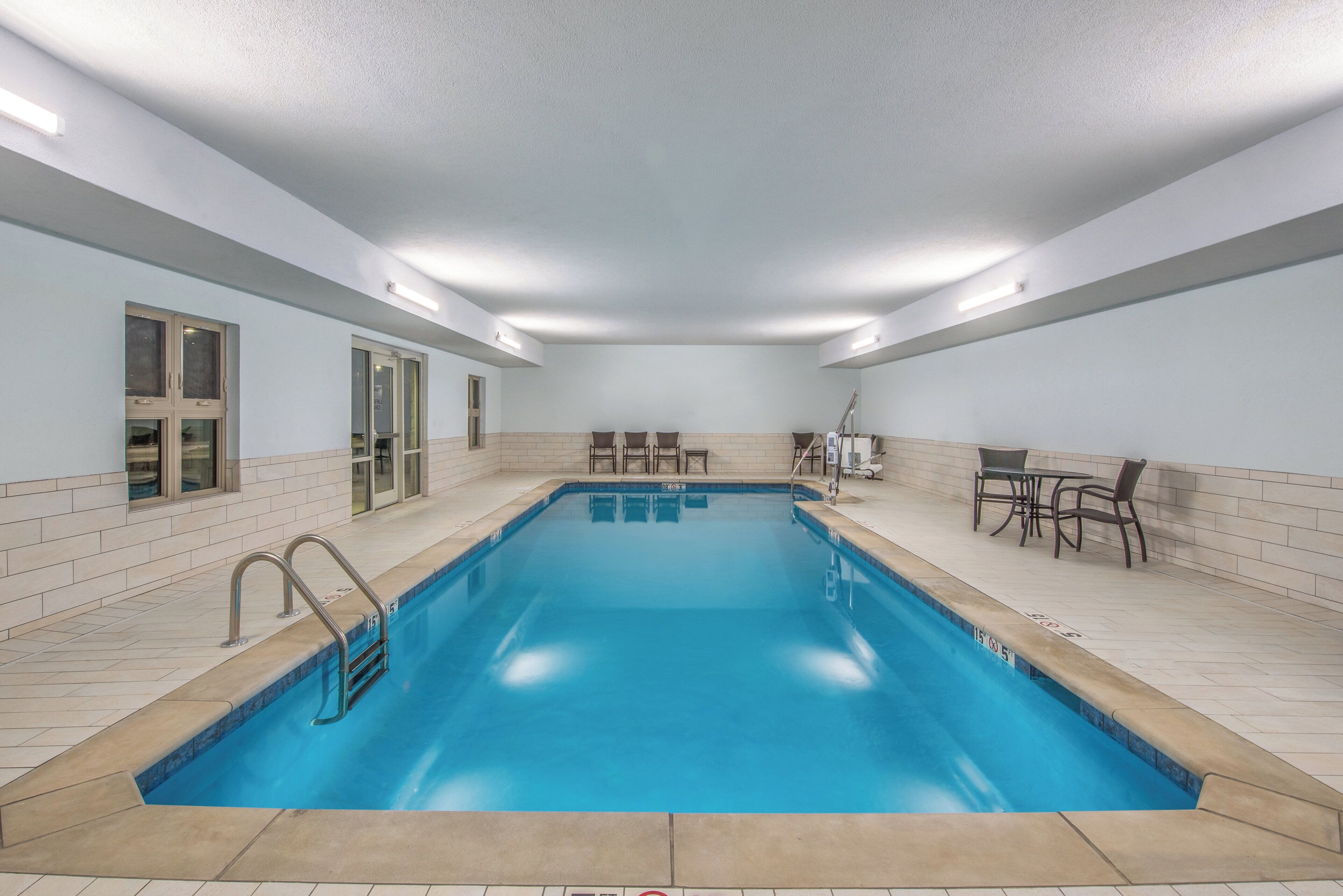 A rectangular indoor swimming pool with clear blue water, surrounded by tiled walls and a ceiling with recessed lighting.