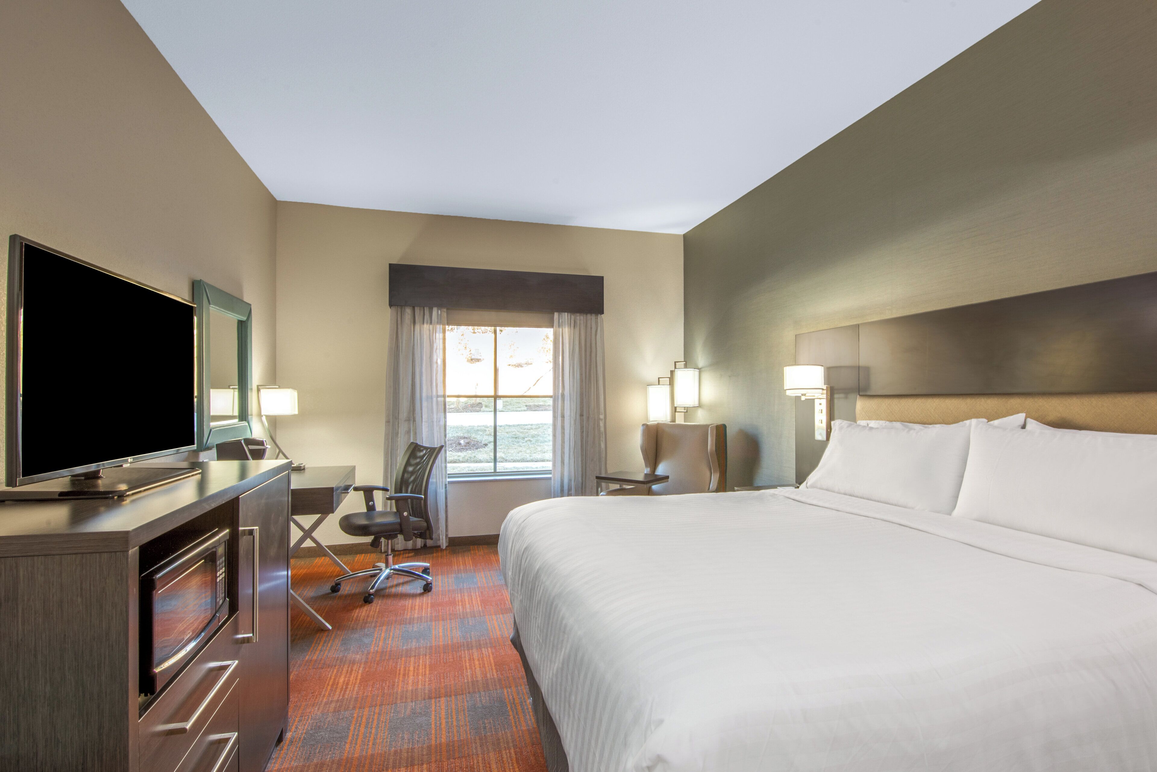 Una habitación de hotel con una cama, un escritorio con televisor, una silla, una ventana con cortinas y una lámpara.
