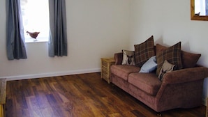 A living room with a wooden floor, a brown sofa with plaid cushions, a small side table, and a window with curtains. - The Grainary Cottages (Scarborough)