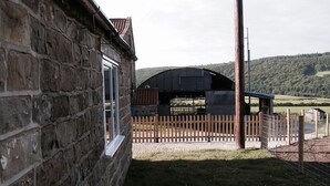 A stone building with a window, a wooden fence, and a barn in the background. - The Grainary Cottages (Scarborough)