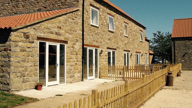 A stone building with multiple windows and a wooden fence. - The Grainary Cottages (Scarborough)
