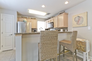 A kitchen with a white door, stainless steel refrigerator, microwave, and wooden cabinets. There are two wicker chairs and a starfish decoration on the wall. - Rain Dancer by South Padre Condo Rentals (South Padre Island)