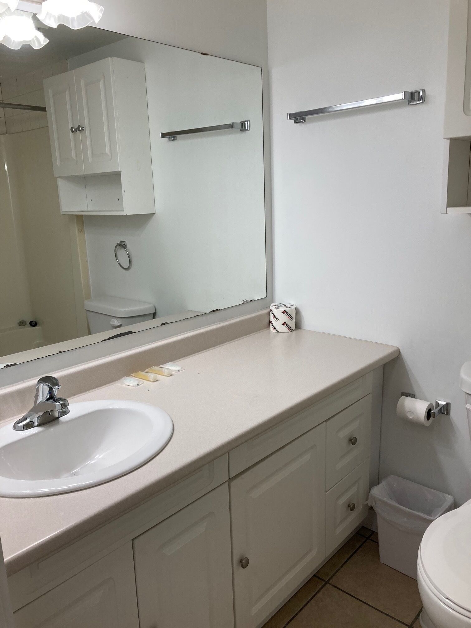 Un baño con un lavamanos blanco, espejo y retrete. Hay un perchero sobre el espejo y un cubo de basura junto al retrete.