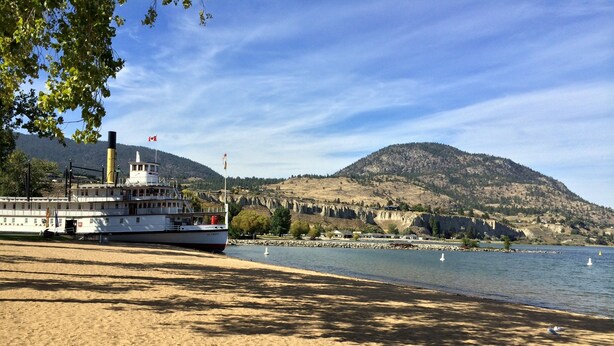 A river with a boat docked on the sandy shore, a mountain in the background, and a clear sky. - Golden Sands Resort Motel (Penticton)