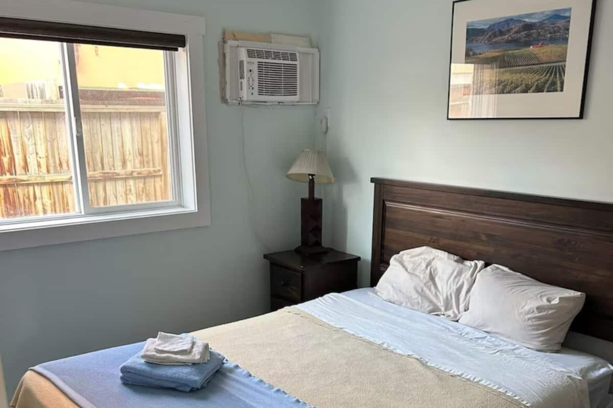Een net opgemaakt bed met een blauw-beige deken, een houten hoofdbord, een bedlampje en een ingelijst schilderij aan de muur.