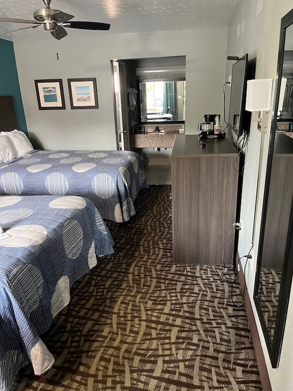 A hotel room with two beds, a ceiling fan, a bathroom with a sink and mirror, and a desk with a lamp.