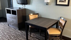 Una camera d'hotel con un tavolo in legno, due sedie, una televisione su un mobile, una cassettiera, una lampada e un quadro appeso al muro.