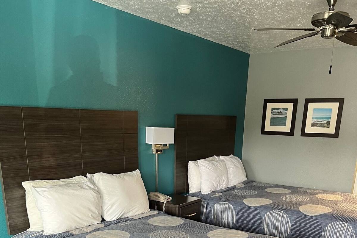 A hotel room with two beds, a desk, a lamp, and a ceiling fan.