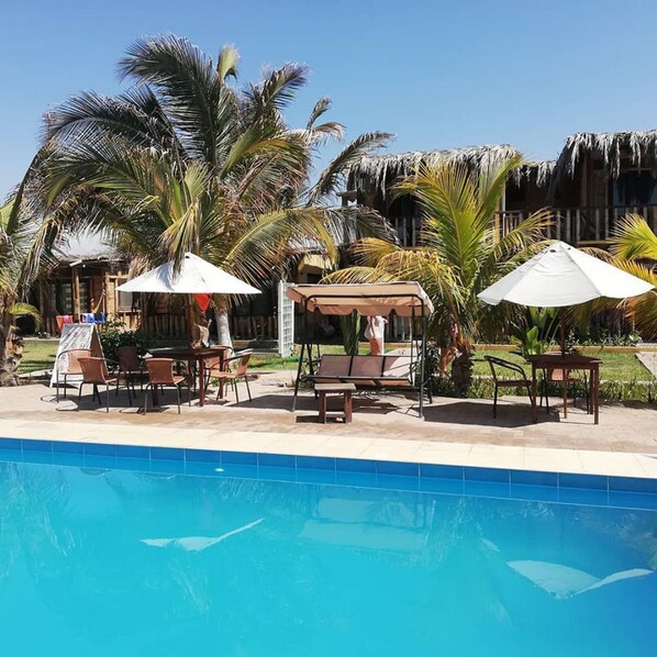 A poolside area with lounge chairs and umbrellas, surrounded by palm trees and a building with a thatched roof. - Punta Pacifico Bungalows Mancora (Máncora)