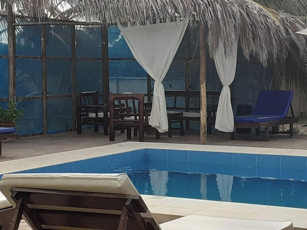 Un área de piscina con piscina azul, sillones de madera para tomar el sol y una estructura de techo de paja.