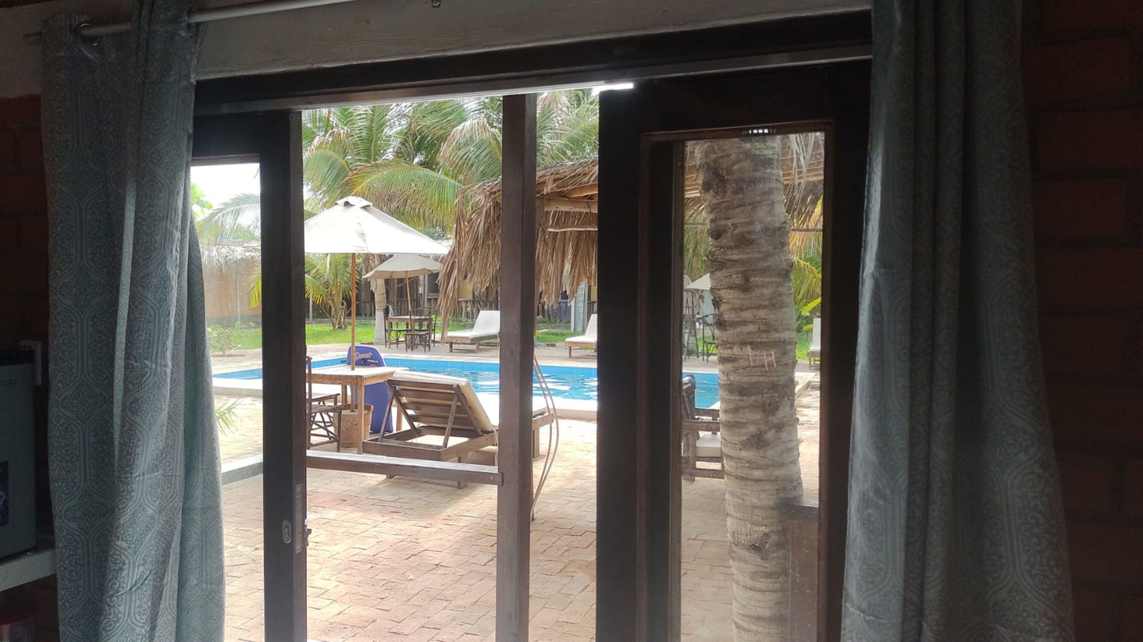 Vista a través de un ventanal de un área de piscina con sillas de descanso, una mesa y una estructura de techo de paja.