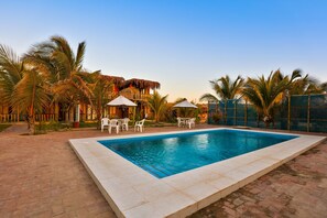 A swimming pool with white chairs and umbrellas, surrounded by palm trees and a building with a thatched roof. - Punta Pacifico Bungalows Mancora (Máncora)