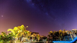 Un resort frente a la playa con palmeras, una piscina y un área de descanso bajo un cielo estrellado.