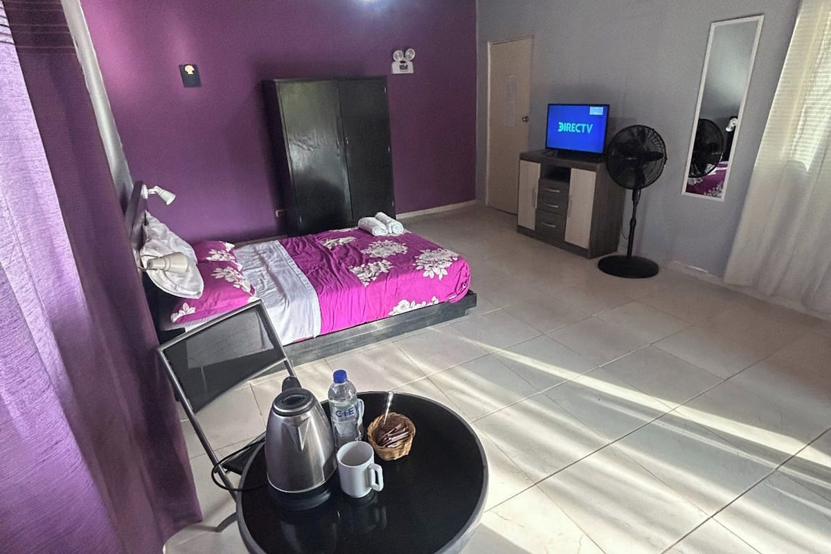 Una habitación con cama, televisión, ventilador y una mesa con tetera y taza.