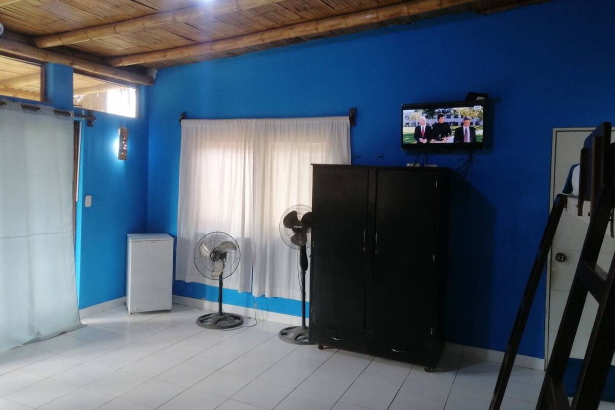 Una habitación con pared azul, techo blanco, armario negro, radiador blanco y cortina blanca, además de un televisor en la pared.
