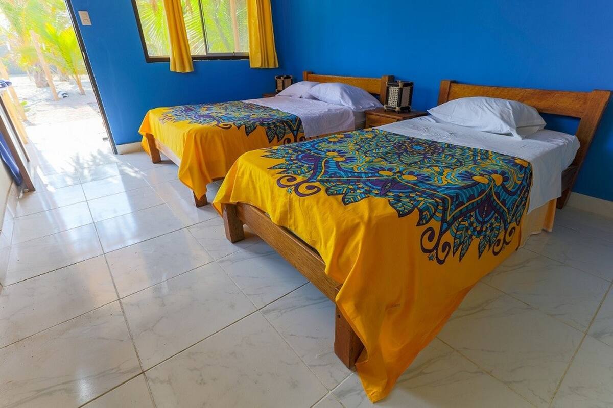 Habitación de hotel con dos camas, cada una con una colcha colorida, un cabecero de madera y una ventana con cortinas amarillas.