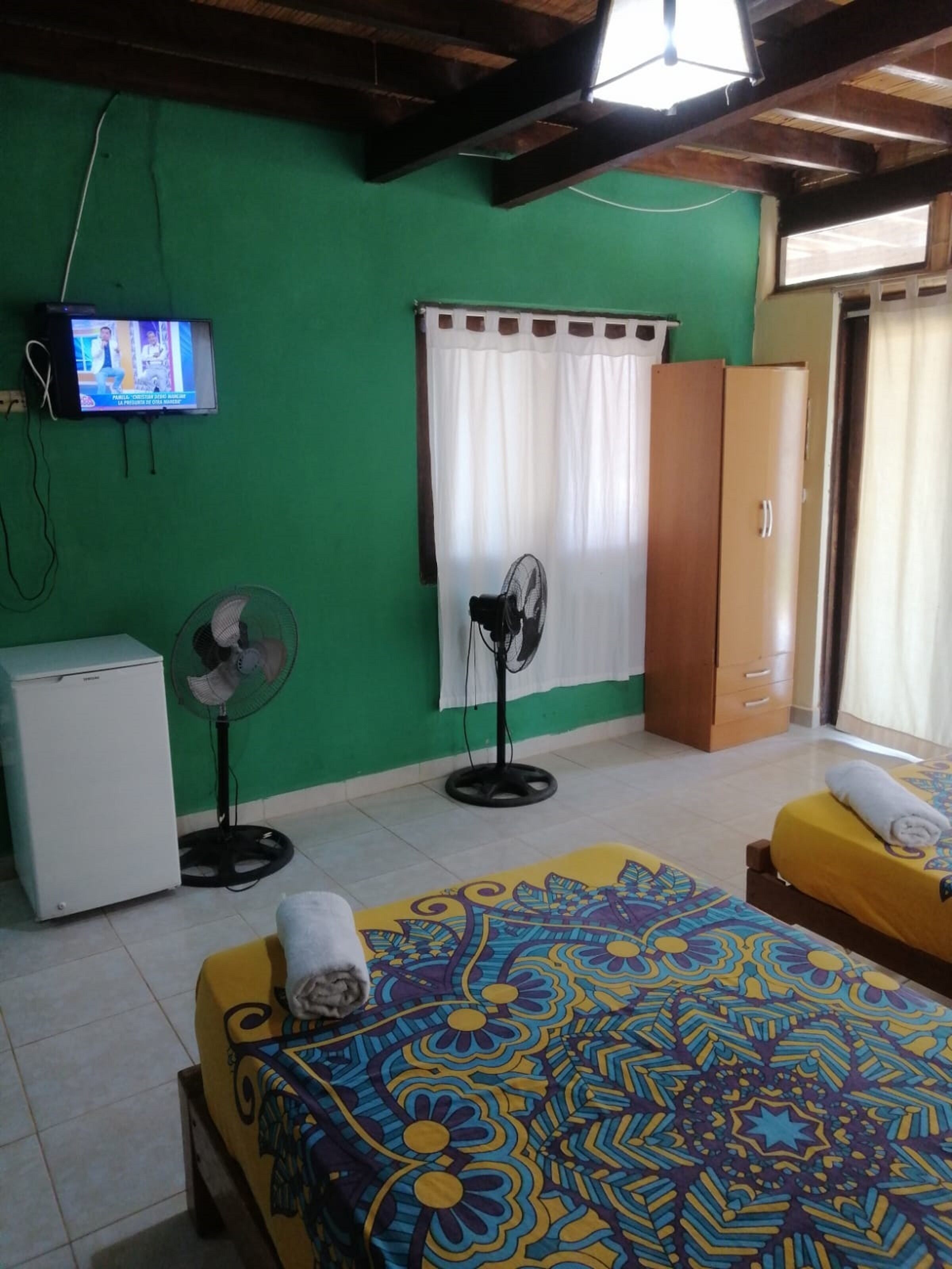 A room with two beds, a small refrigerator, a TV on the wall, a fan, and a wardrobe.