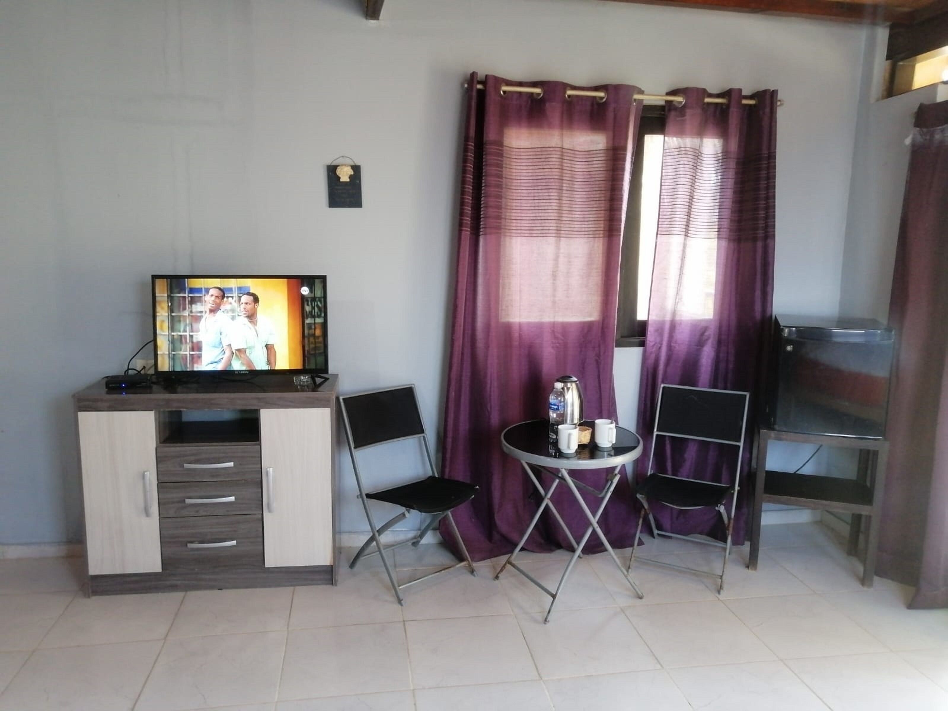 A small room with a TV on a wooden cabinet, two chairs, a small table with a kettle and cups, and purple curtains.