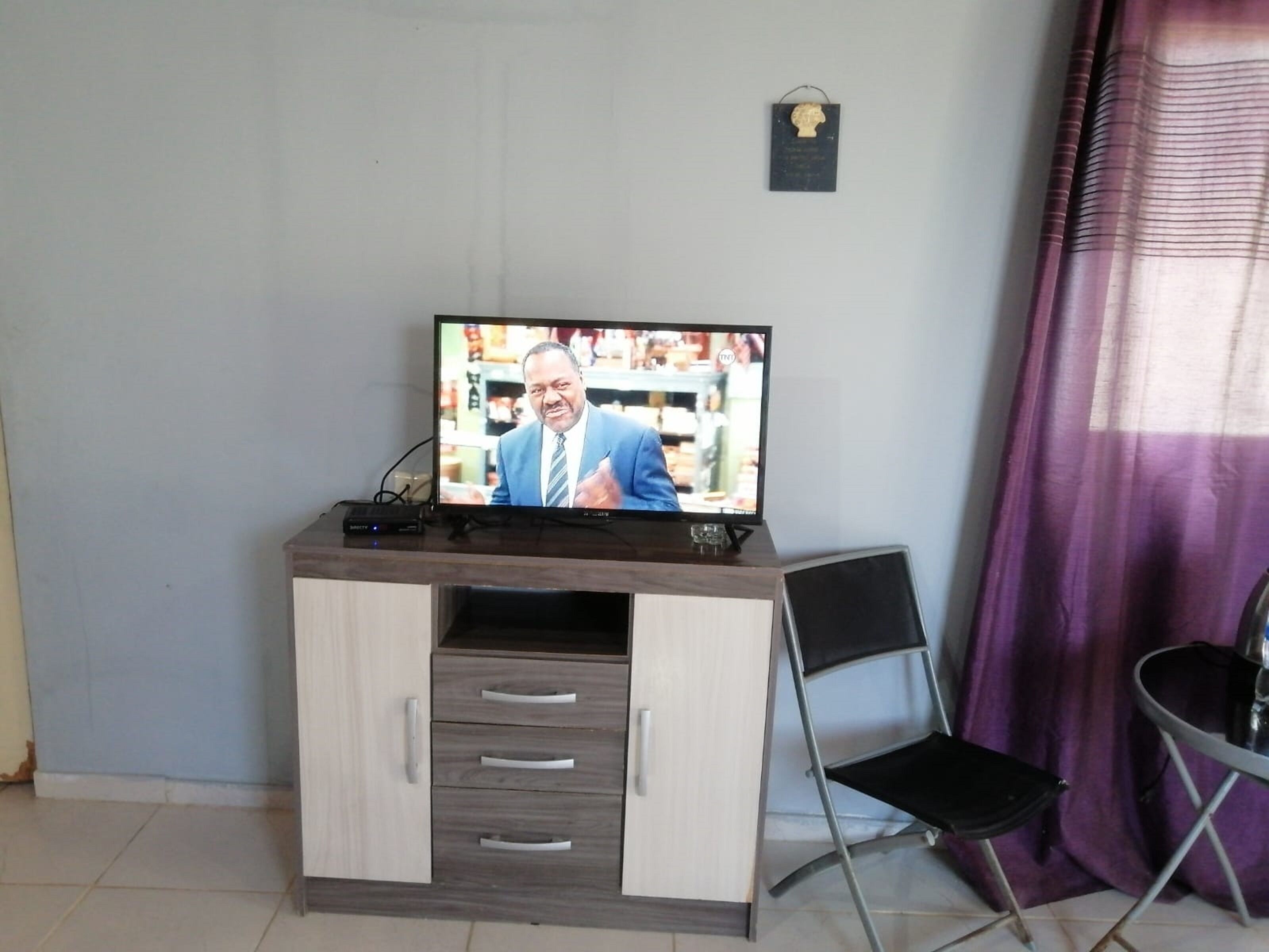 A television on a wooden cabinet with drawers, a folding chair, and a purple curtain in the background.