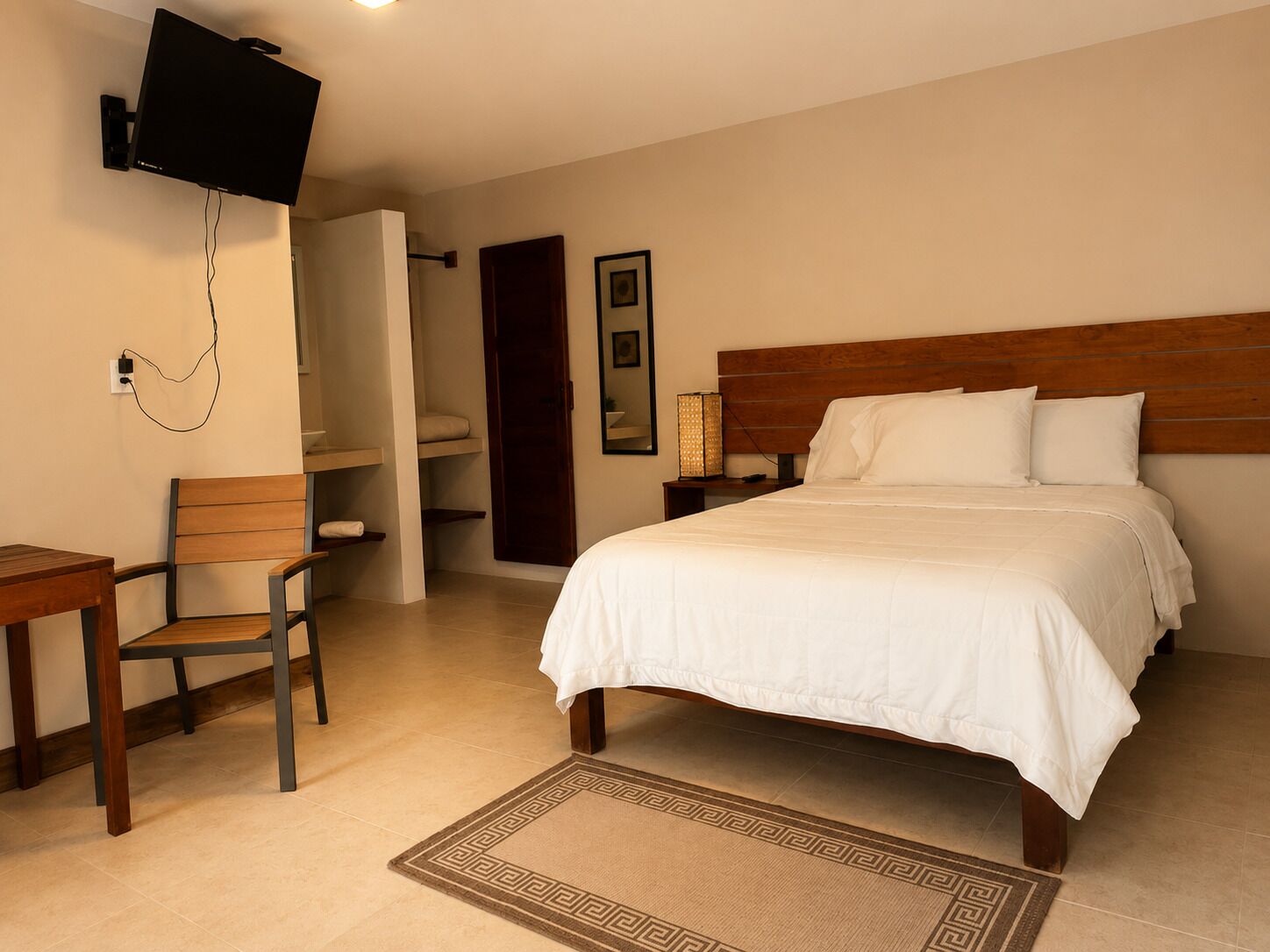 Habitación de hotel con una cama, un escritorio, una silla, un televisor montado en la pared y una mesita de noche con una lámpara.