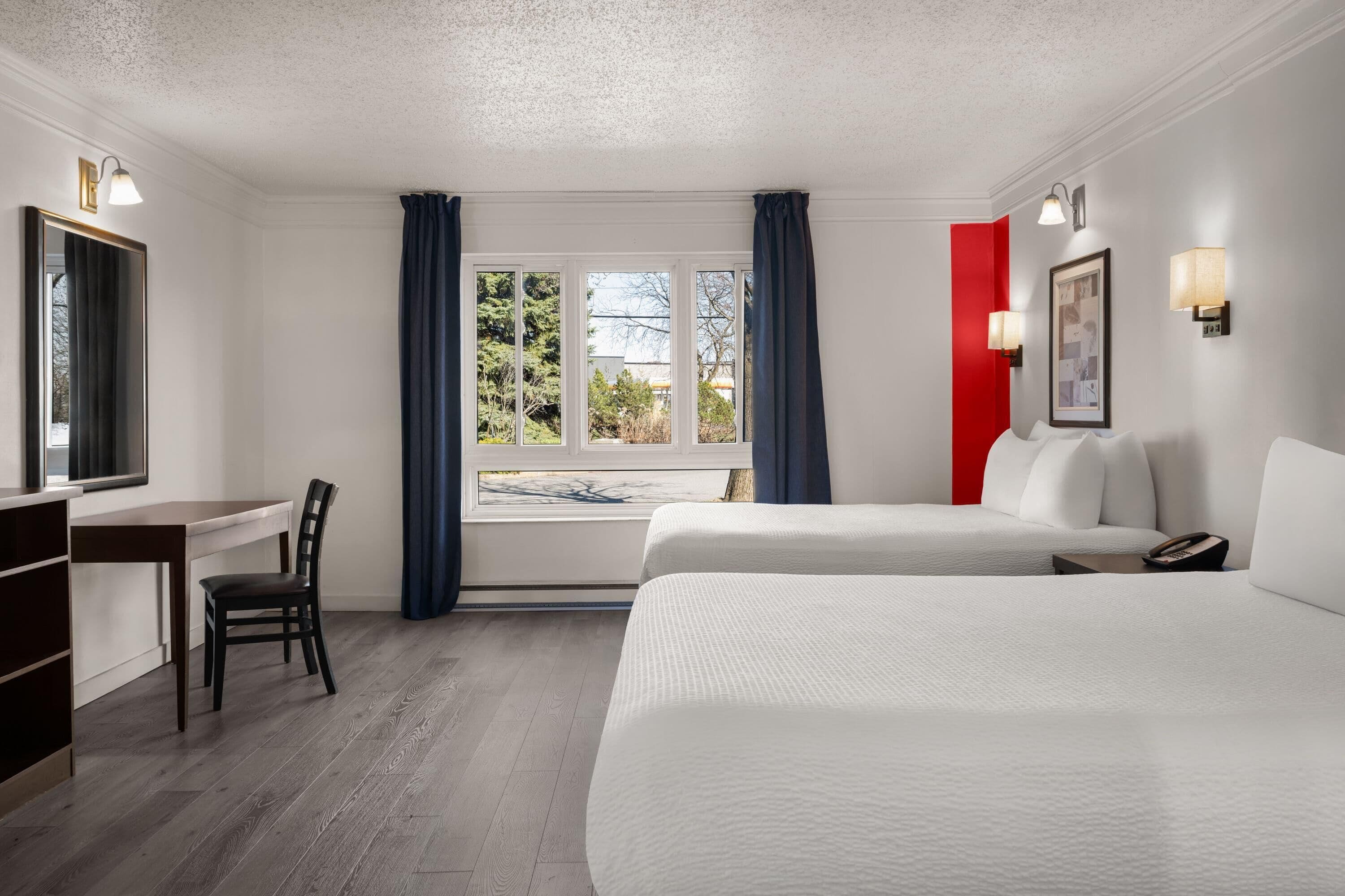 A hotel room with a desk, two beds, a chair, a window with curtains, and wall-mounted lights.