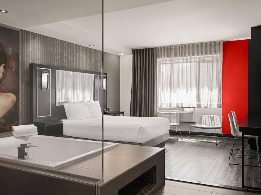 A modern hotel room with a large bed, a separate seating area, a large mirror, and a bathtub.