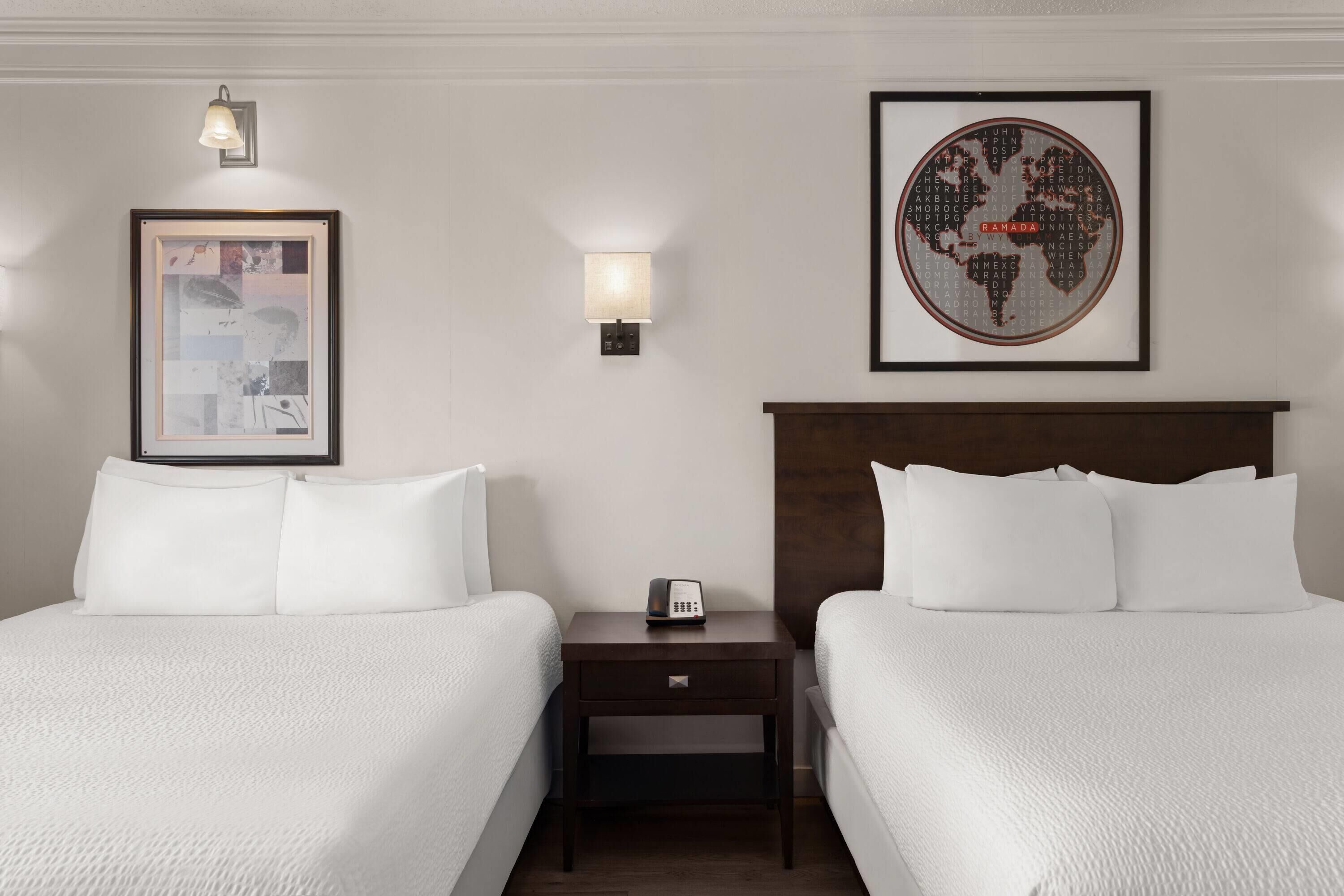 A hotel room with two beds, a nightstand with a phone, a framed abstract artwork, a wall-mounted lamp, and a framed world map.