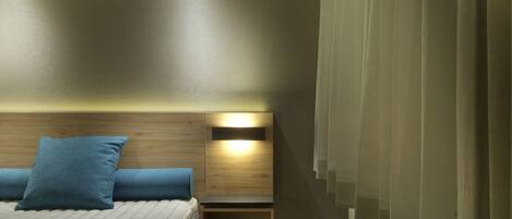 A modern hotel room with a bed, a bedside table, a wall-mounted shelf, and a window with curtains.