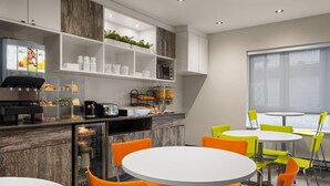A modern kitchen with a coffee machine, shelves with cups, and a refrigerator. There are orange and yellow chairs around a white table. - Ramada by Wyndham Laval (Laval)