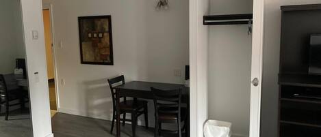 A room with a dining table and chairs, a small white trash bin, and a painting on the wall.