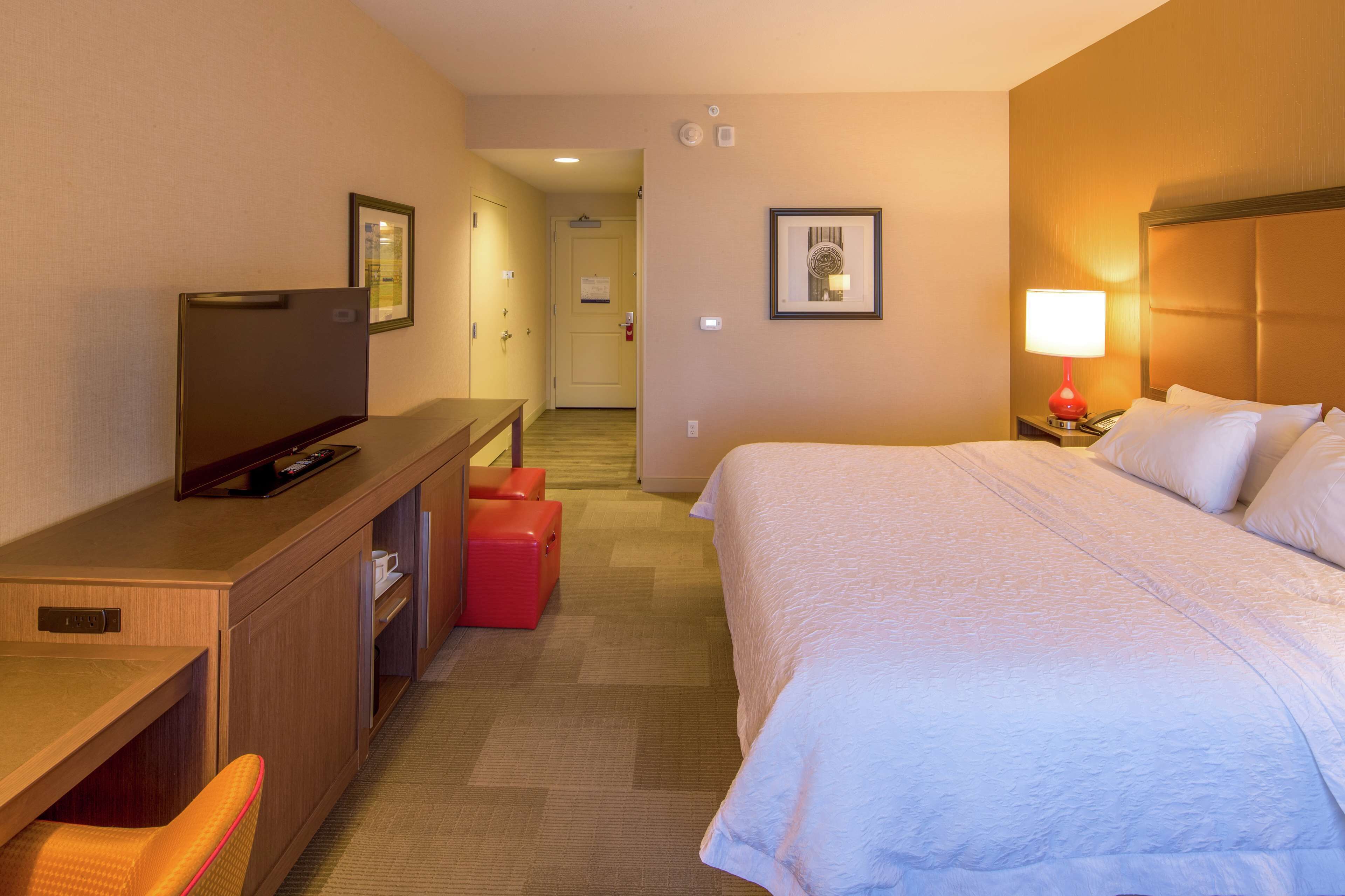 Quarto de hotel com uma cama, uma escrivaninha com televisão, um banquinho vermelho e uma mesa de cabeceira com abajur.