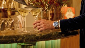 A bar area with a person holding a martini glass, a watch on their wrist, and a vase with red flowers.