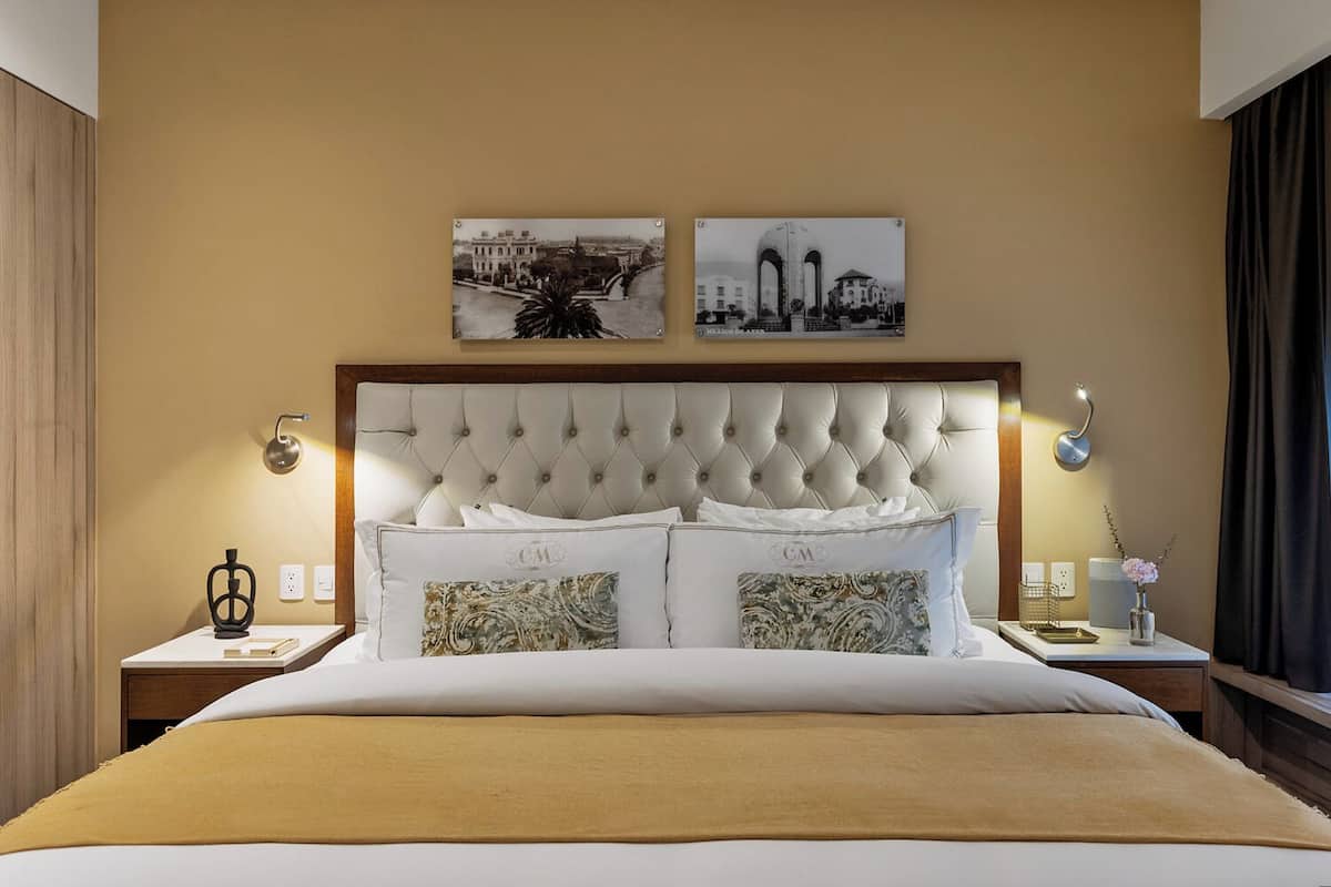 A hotel room with a tufted headboard, a large bed with white linens, two decorative pillows, a bedside table with a lamp, and two framed pictures on the wall.